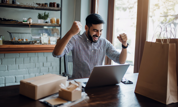 Empresário comemorando resultados após criar página de vendas de sucesso no notebook, com embalagens de produtos ao redor.